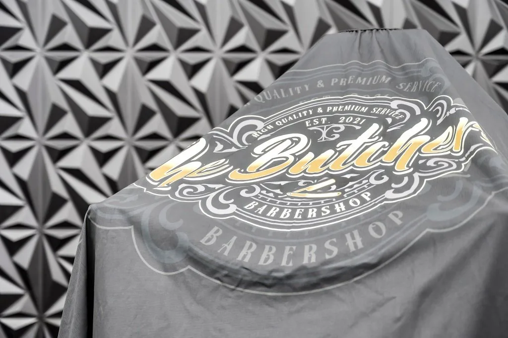 The Butchers Barbershop cape with the gold crest draped over the chair — High Quality & Premium Service, Est. 2021