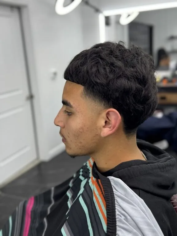 Low fade with a textured curly top and a crisp temple lineup — young client in profile wearing the striped cape