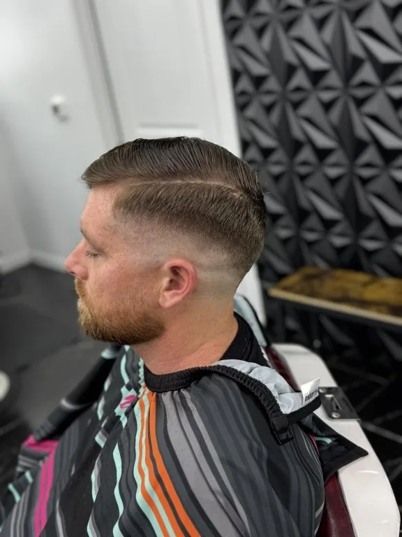 Skin fade with a razor hard part and a full groomed beard — finished cut shown in profile in the chair